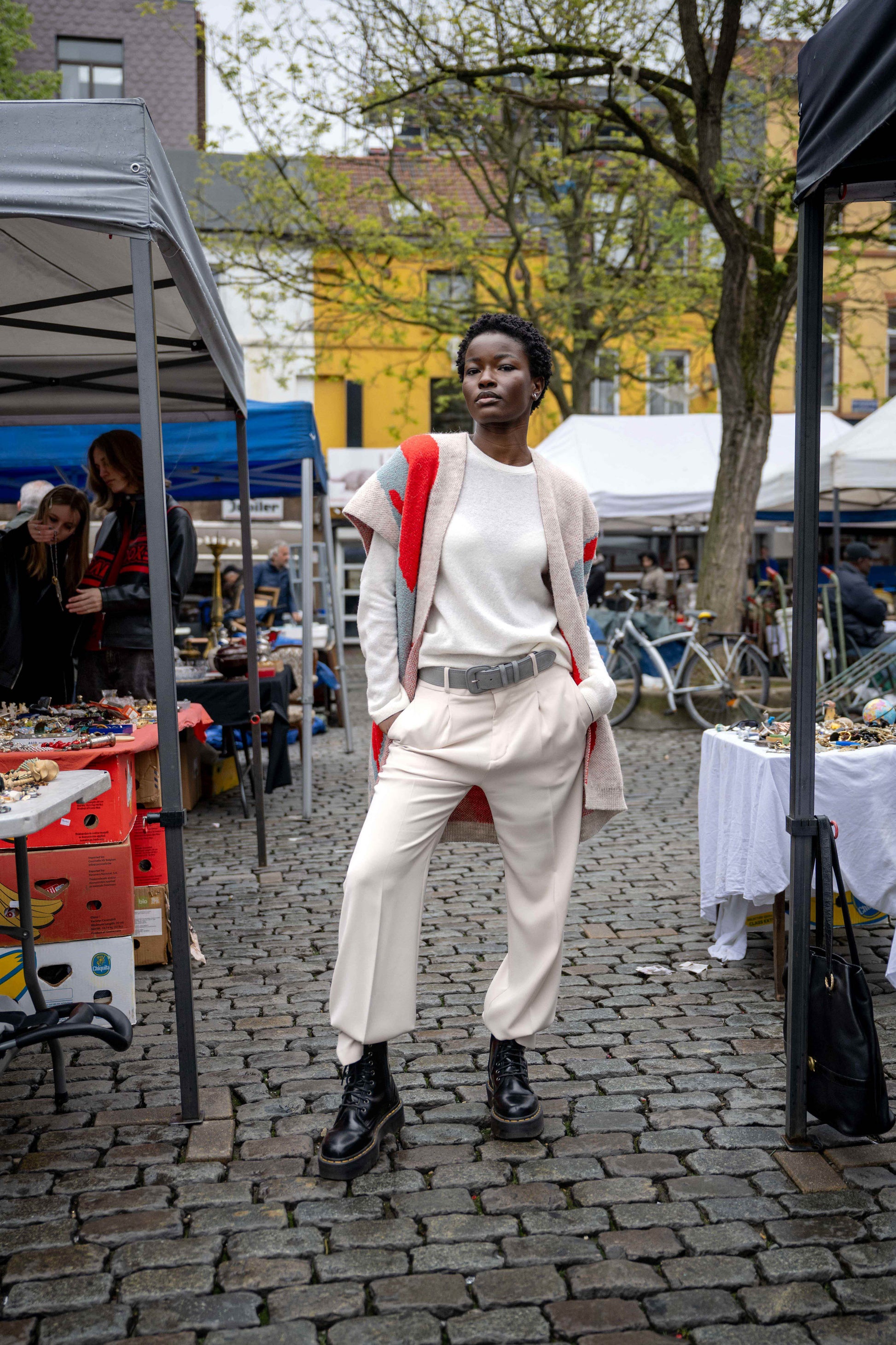 Confident girl standing between two tents, wearing white pants with black combat boots, a white wool sweater, and a long Ohara coat with red and blue accents.