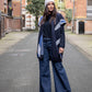 Girl standing between tall buildings, wearing jeans, a beautiful long blue coat, a blue sweater, and a gray beanie.