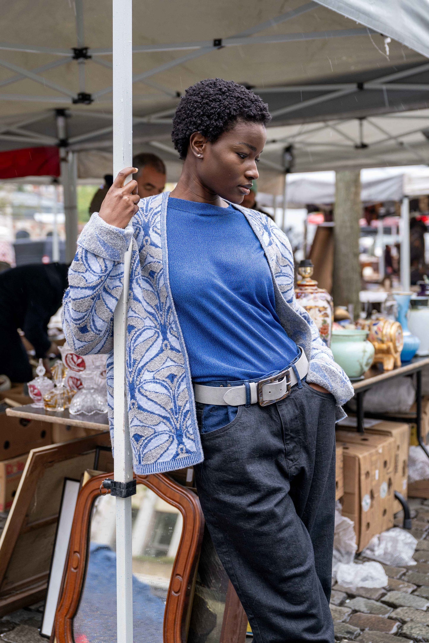 Black girl leaning against a tent, wearing a blue sweater with a special gray and blue jacquard pattern, a gray belt, and cool pants.