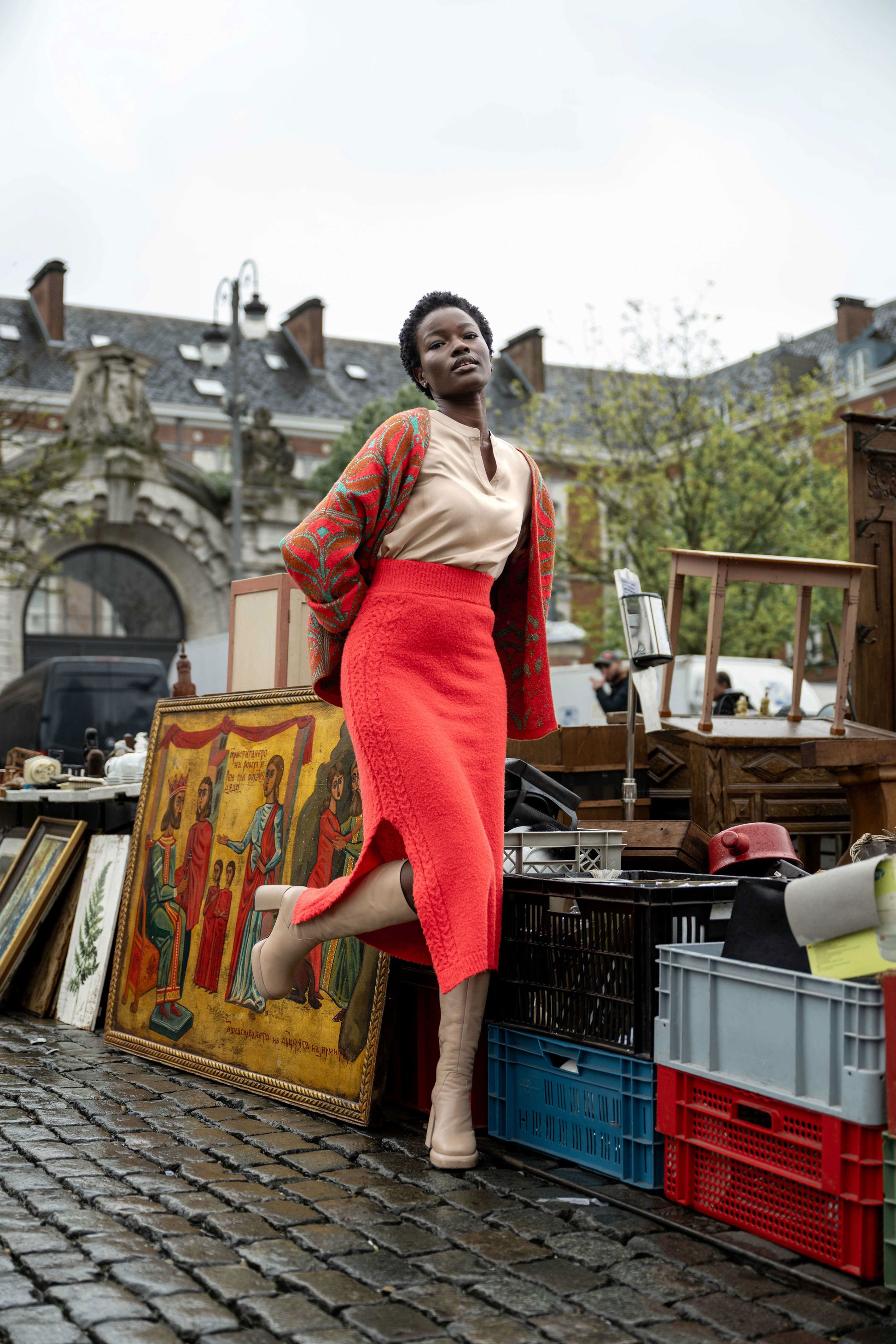 Woman leaning against a gold painting, wearing a beautiful red form-fitting skirt, a beige blouse, and a red and green jacquard vest.