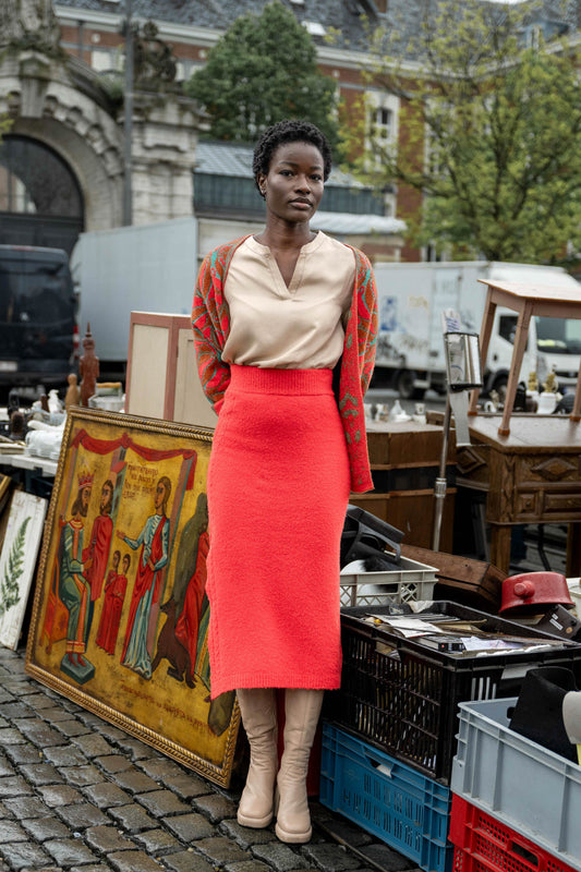 Woman leaning against a gold painting, wearing a beautiful red form-fitting skirt, a beige blouse, and a red and green jacquard vest.