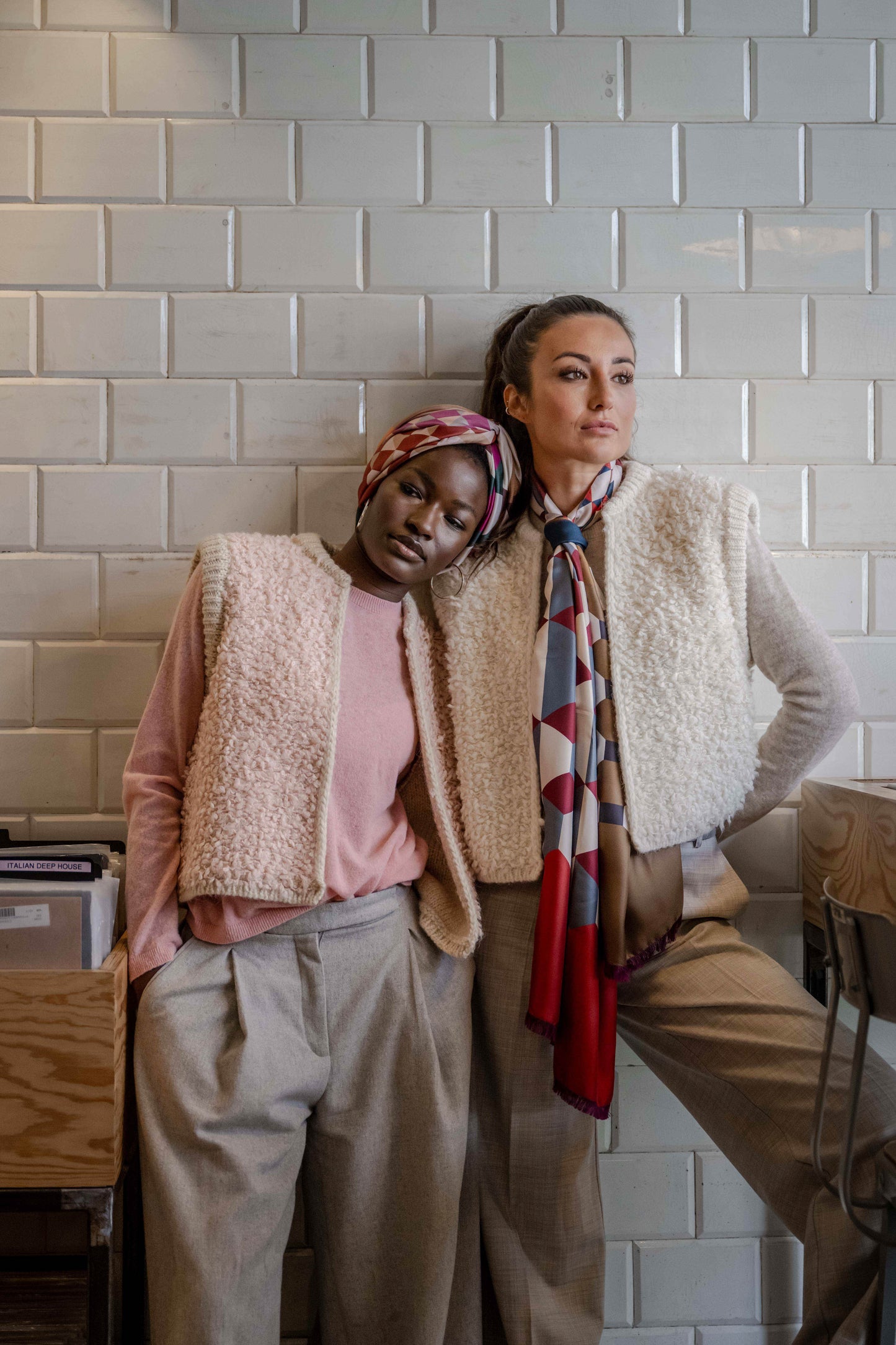 Two girls leaning against a wall with their heads touching, wearing similar outfits: both have silk scarves, beige fluffy vests over wool sweaters