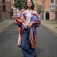 Woman standing between tall buildings, wearing a silk kimono with a graphic print in purple and orange shades, long jeans, and cowboy boots.