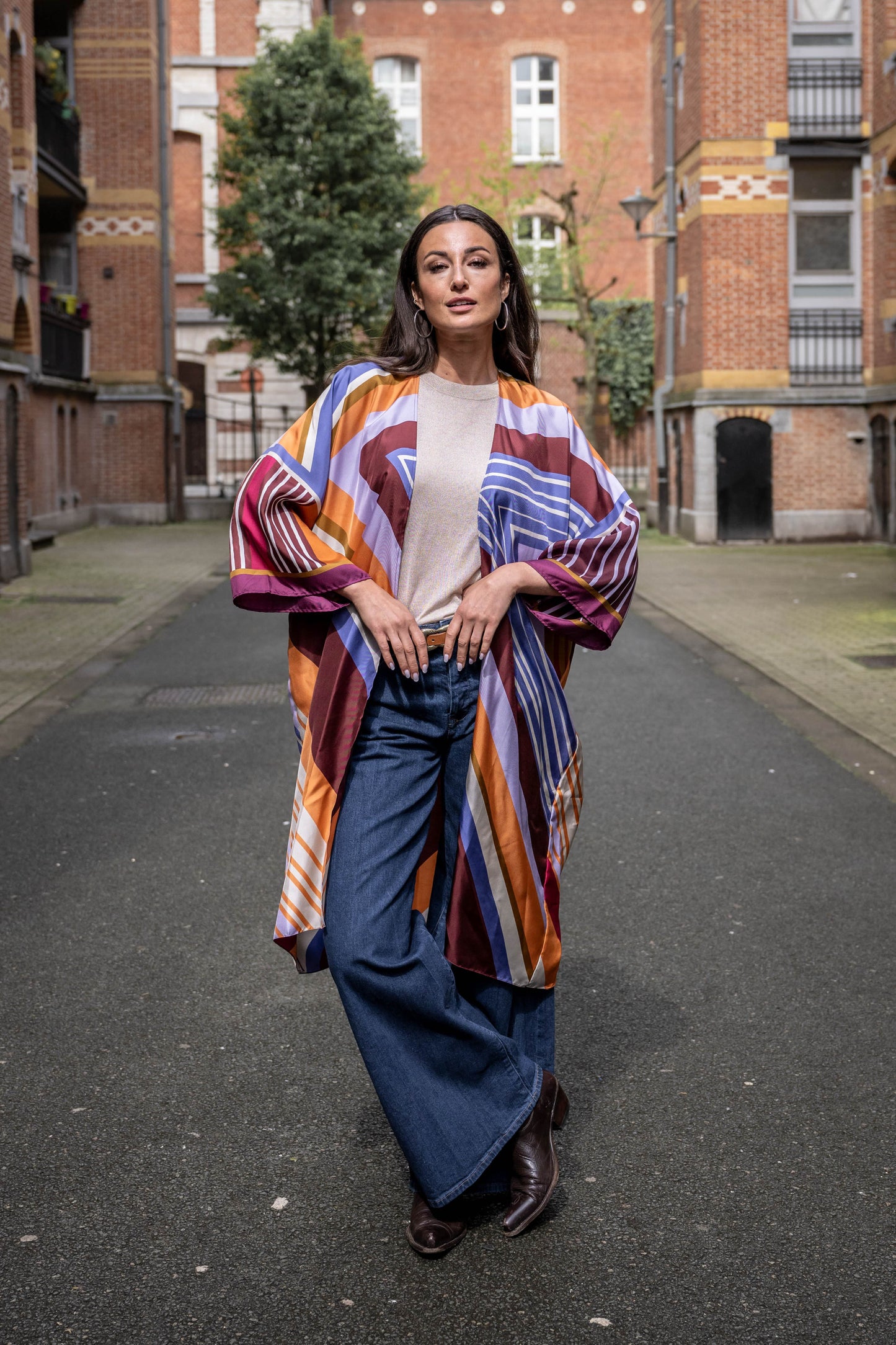 Woman standing between tall buildings, wearing a silk kimono with a graphic print in purple and orange shades, long jeans, and cowboy boots.