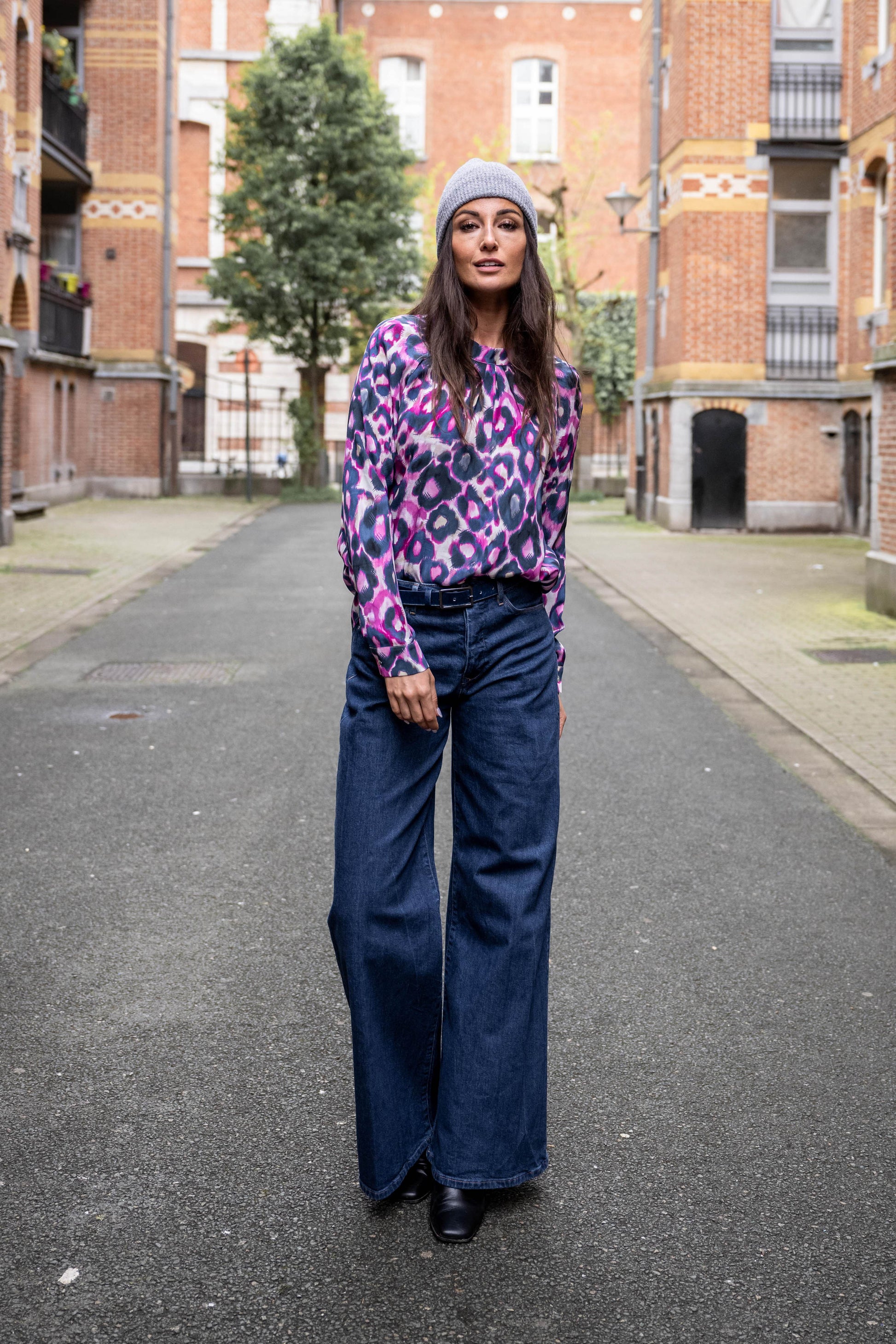 girl standing between big buildings, wearing a beanie, silk blouse, jeans and leather belt