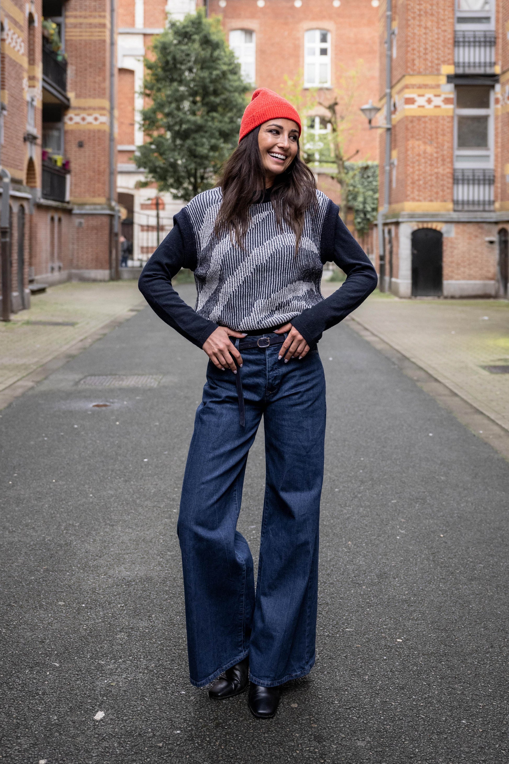 Women is wearing long jeans with leather belt, cardigan with artwork in dark blue and wears a beanie in red